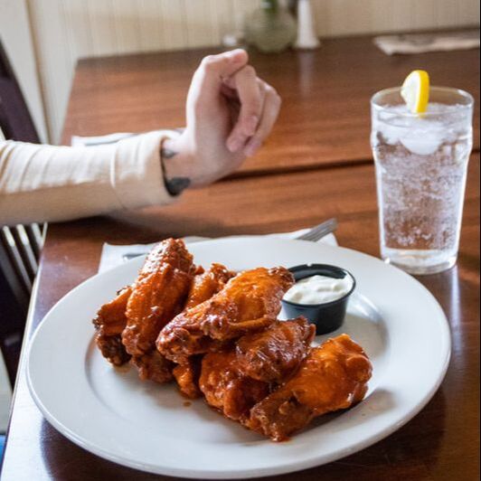 Picture of a plate full of jumbo wings and a persons arm in the background with a glass of water